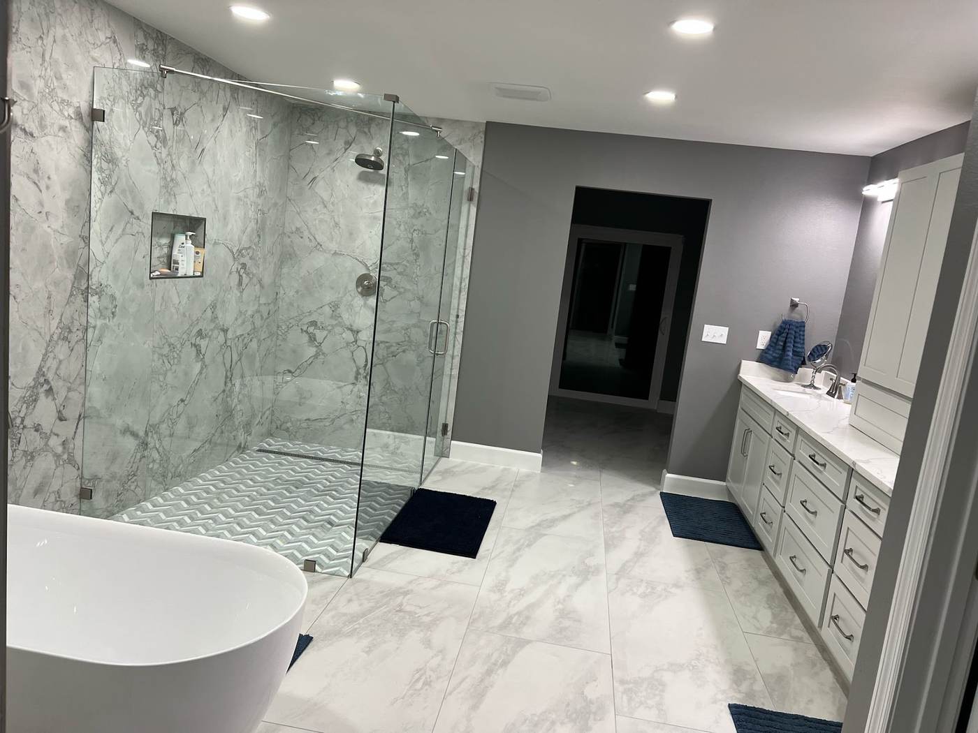 Marble-look wall tile and a herringbone mosaic shower floor behind glass, with a freestanding tub and large-format floor tile.